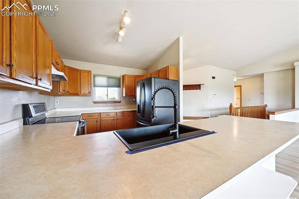 Image 11 of 50: Kitchen with brown cabinetry, vaulted ceiling, light countertops, stainless