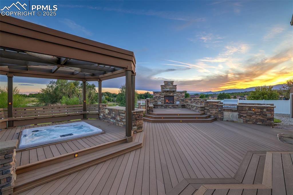 Image 43 of 50: Deck at dusk with exterior kitchen, an outdoor stone fireplace, and ceiling