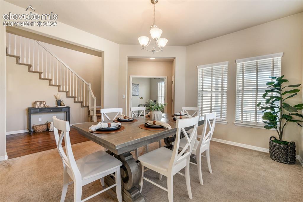Image 5 of 50: Carpeted dining area with a chandelier and two windows featuring blinds
