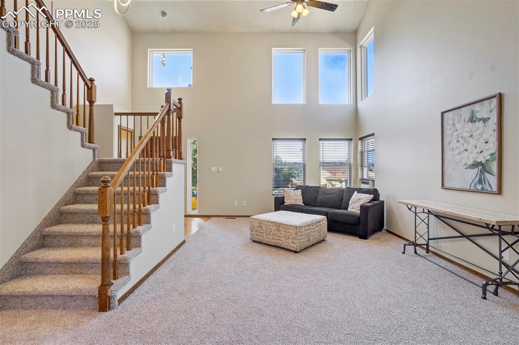 Image 7 of 50: Carpeted living room featuring a ceiling fan, a towering ceiling, and stair