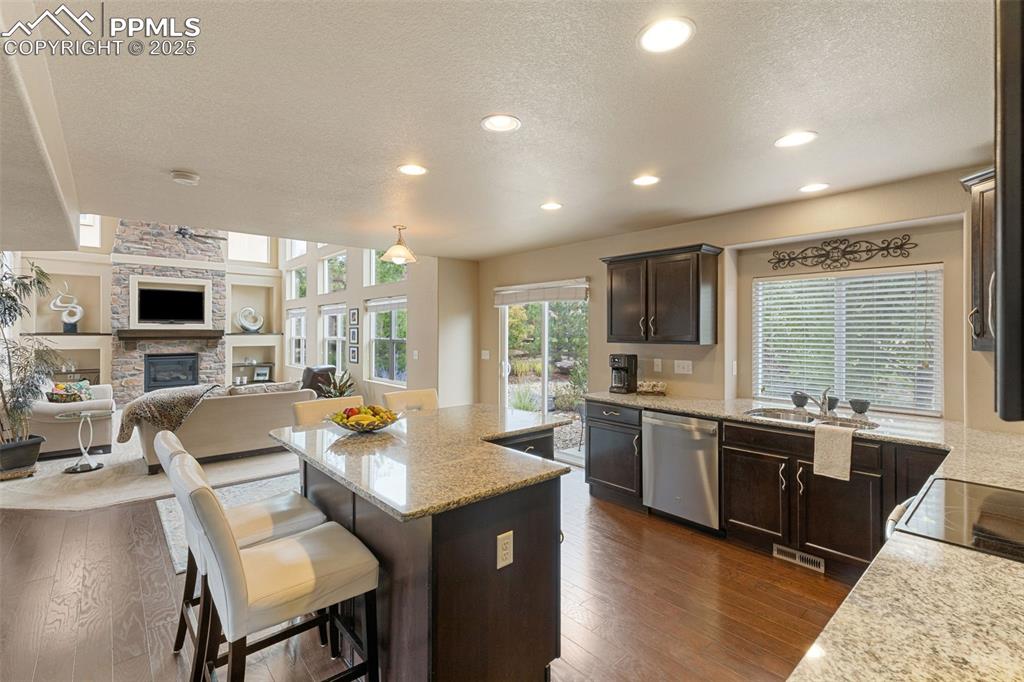 Image 11 of 50: Kitchen with dark brown cabinets, a stone fireplace, open floor plan, a tex
