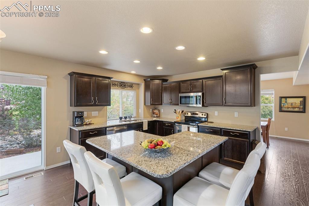 Image 12 of 50: Kitchen featuring dark brown cabinetry, a kitchen bar, appliances with stai
