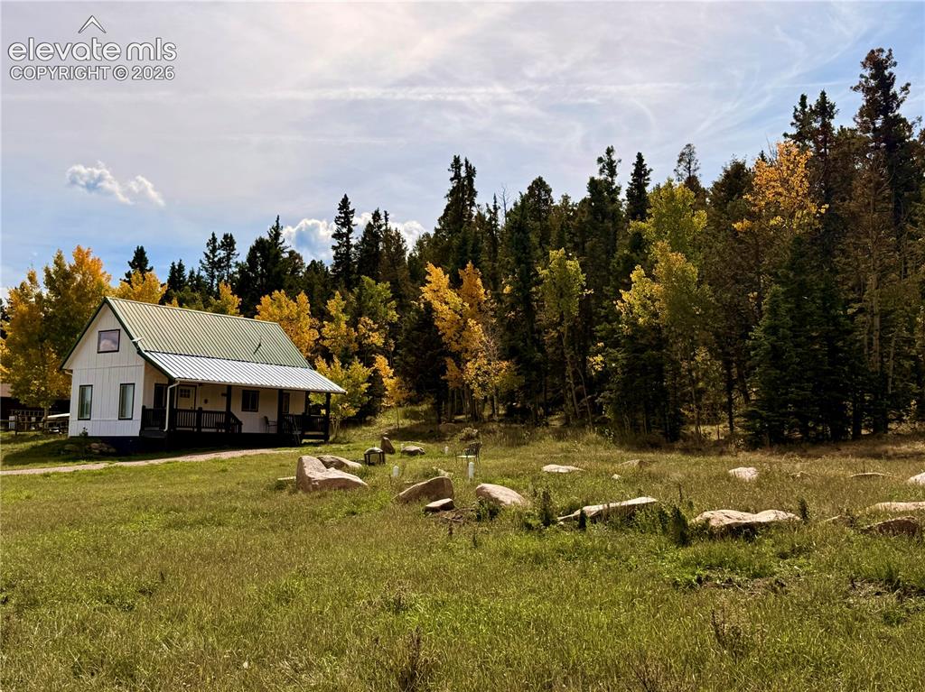 Image 2 of 43: White exterior siding, green metal roof, covered porch, surrounding mature 