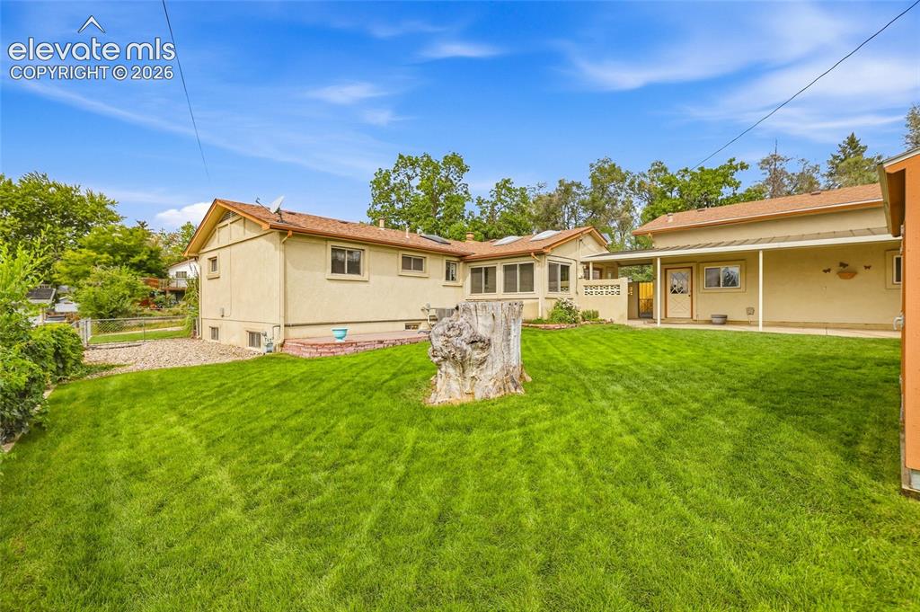 Image 33 of 43: Expansive grassy lawn with a large tree stump, stucco exterior, and covered