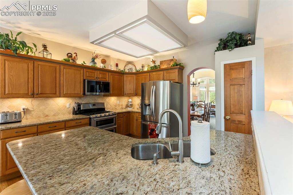 Image 19 of 50: Kitchen featuring arched walkways, brown cabinetry, appliances with stainle