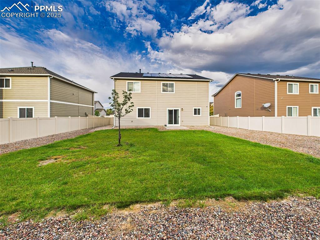 Image 42 of 50: Oversized fenced backyard with green lawn, rock landscaping, and solar pane