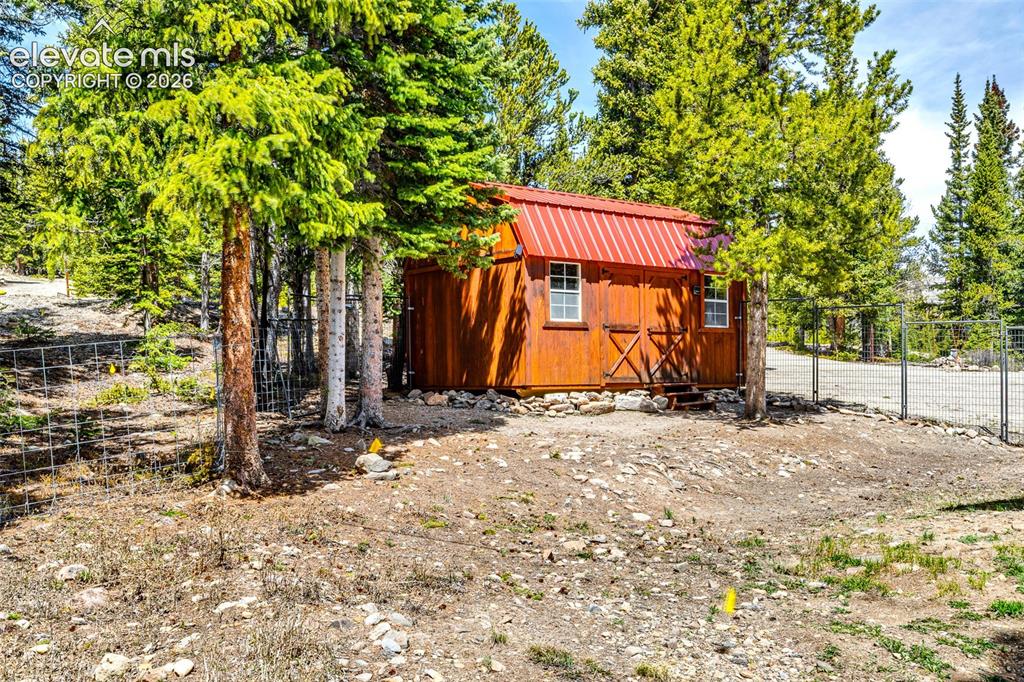 Image 30 of 37: Wood-sided outbuilding with a red metal roof and white-framed windows, surr
