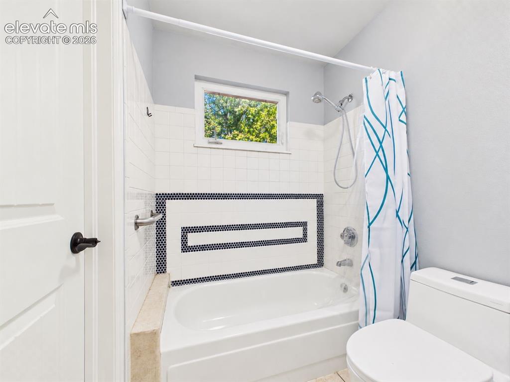 Image 28 of 50: Tub and shower surround features white tile accented by a geometric dark mo