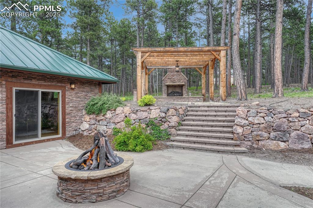 Image 42 of 50: Stone stairs lead to a pergola with an outdoor grill and gas log fireplace