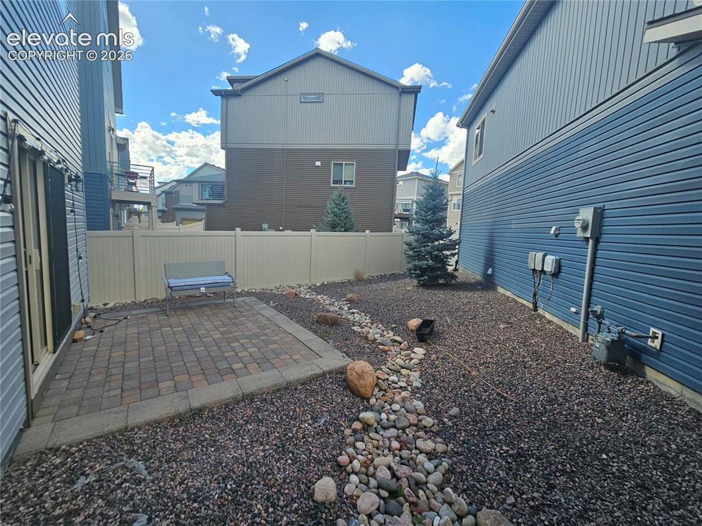 Image 22 of 23: Paved patio area, rock landscaping, mature evergreen trees, and a vinyl pri