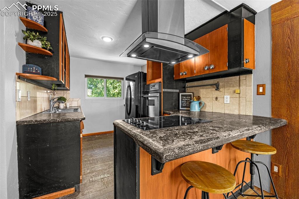 Image 14 of 42: Kitchen with island exhaust hood, black appliances, open shelves, backsplas