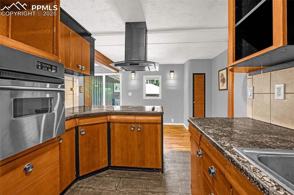 Image 16 of 42: Kitchen with oven, island exhaust hood, black electric stovetop, dark wood-