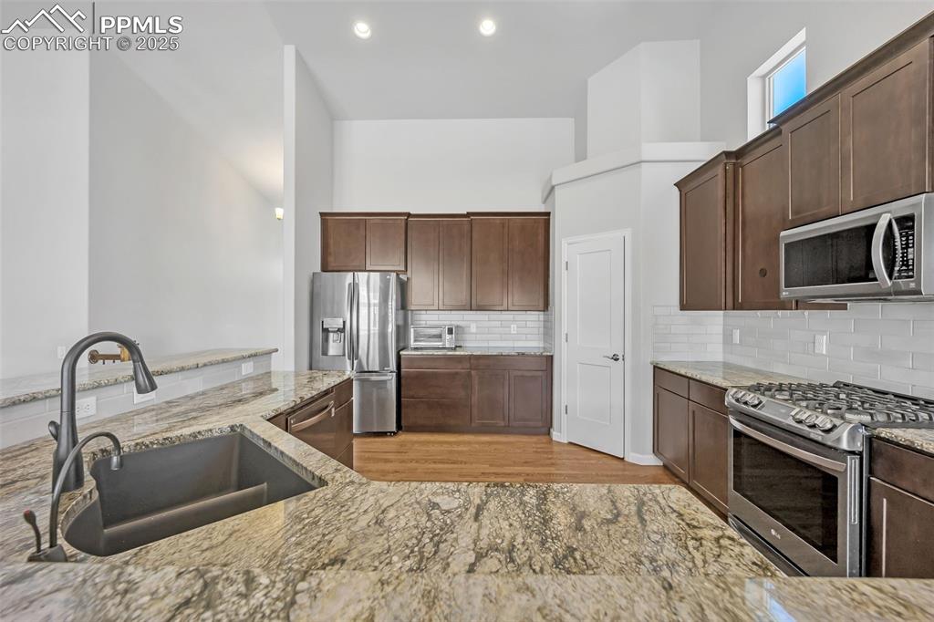 Image 8 of 45: Kitchen with dark brown cabinetry, light stone countertops, appliances with
