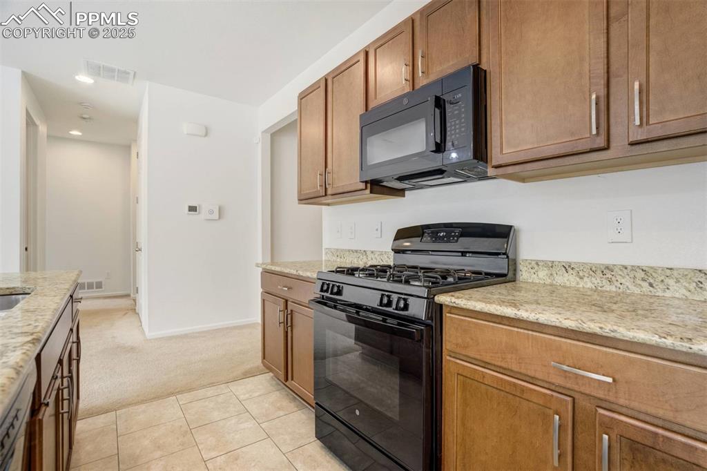 Image 10 of 36: Kitchen with black appliances, light carpet, brown cabinetry, light stone c