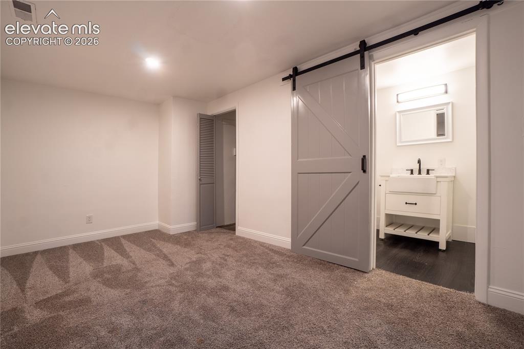 Image 23 of 33: Spacious room with gray patterned carpet, smooth white walls, and bright ov