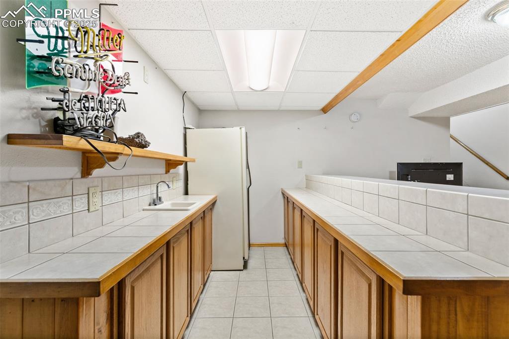 Image 30 of 36: Kitchen featuring tile countertops, brown cabinetry, a paneled ceiling, ope