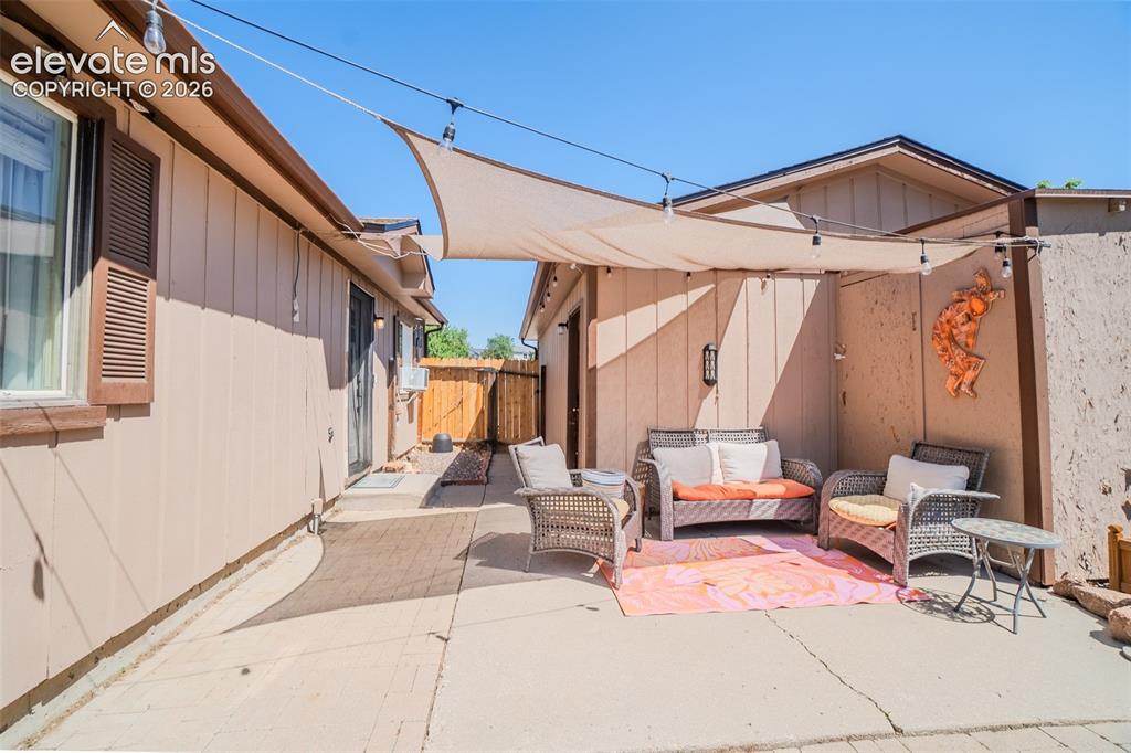 Image 24 of 31: Paved patio area featuring a sun shade and string lighting, bordered by ver