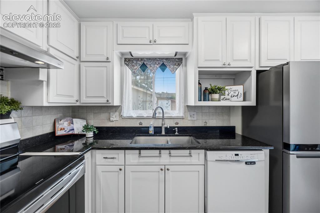 Image 6 of 25: Kitchen featuring white cabinetry, dark countertops, a double basin sink, a