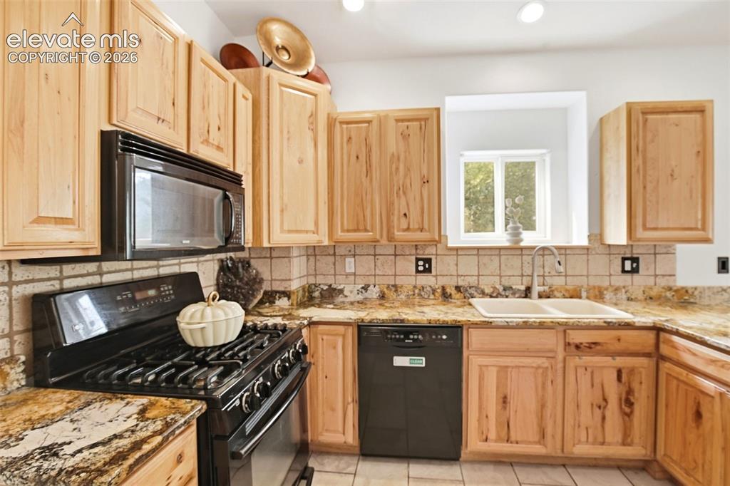 Image 29 of 49: Light wood cabinetry with black appliances, tile backsplash, and stone coun
