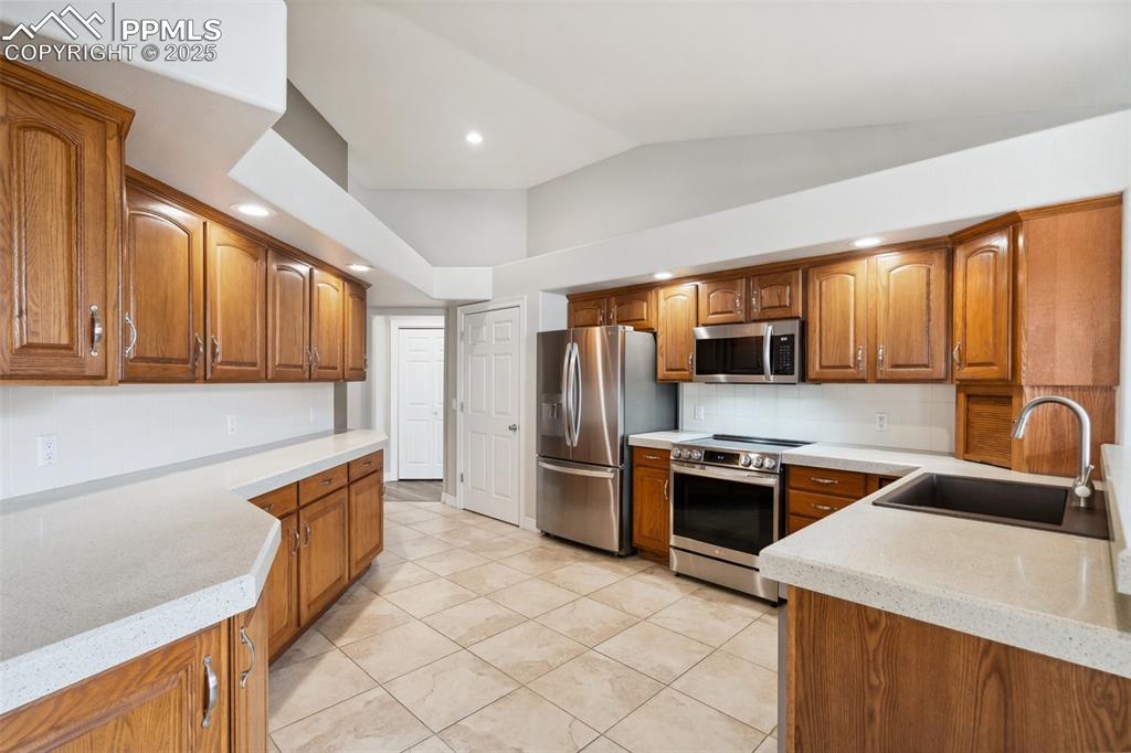 Image 12 of 49: Kitchen with vaulted ceiling, backsplash, a sink, brown cabinetry, and stai