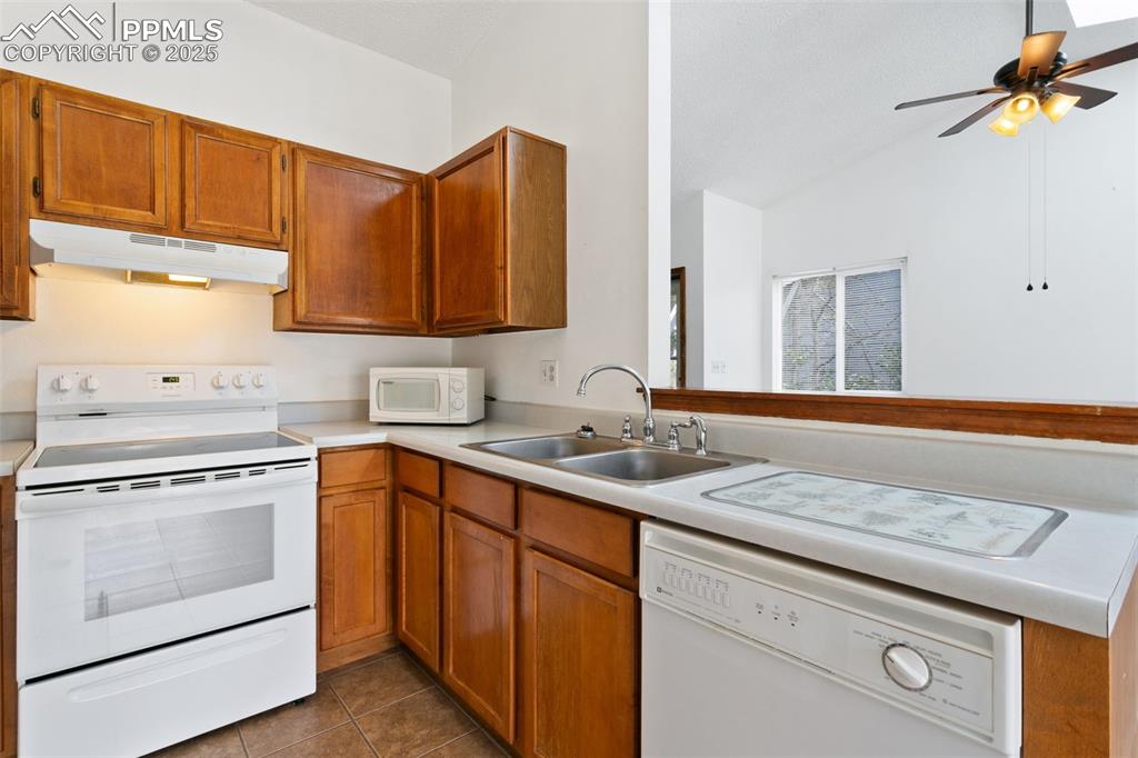 Image 13 of 24: Kitchen with white appliances, brown cabinetry, light countertops, under ca