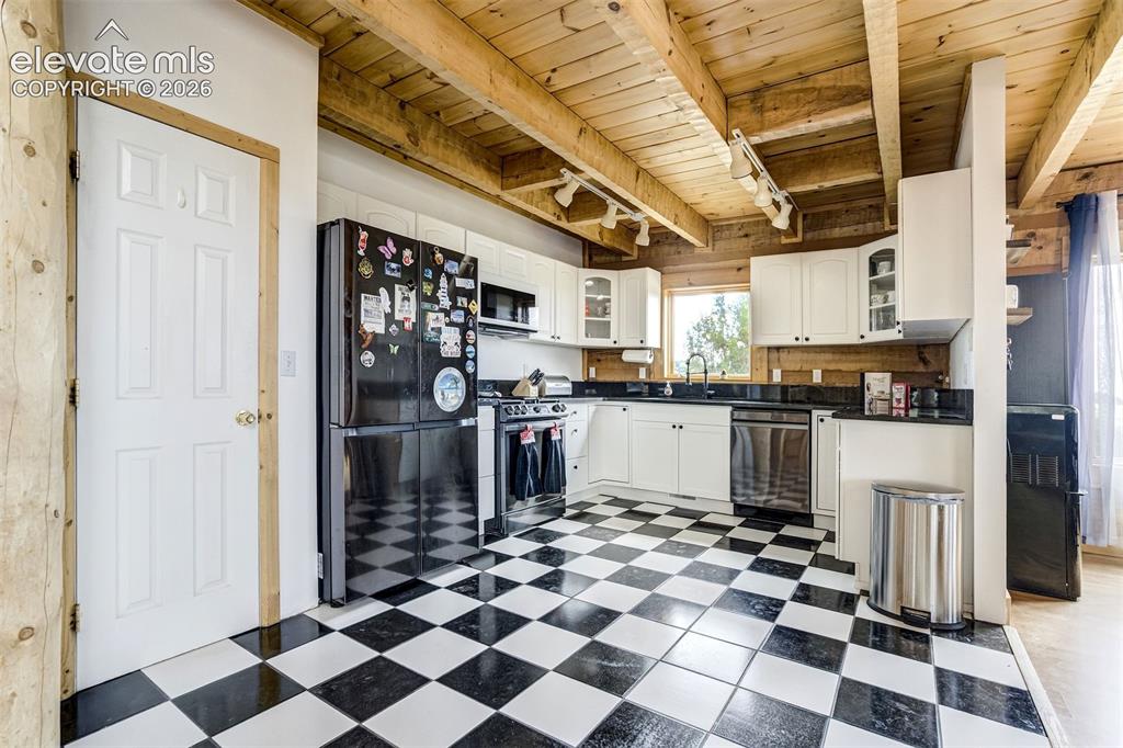 Image 15 of 50: Kitchen featuring exposed wood beam ceilings, white cabinetry, black counte