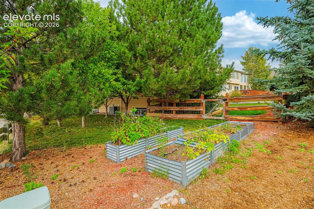 Image 38 of 50: Raised garden beds with plantings, mature trees, and a wooden fence with ga