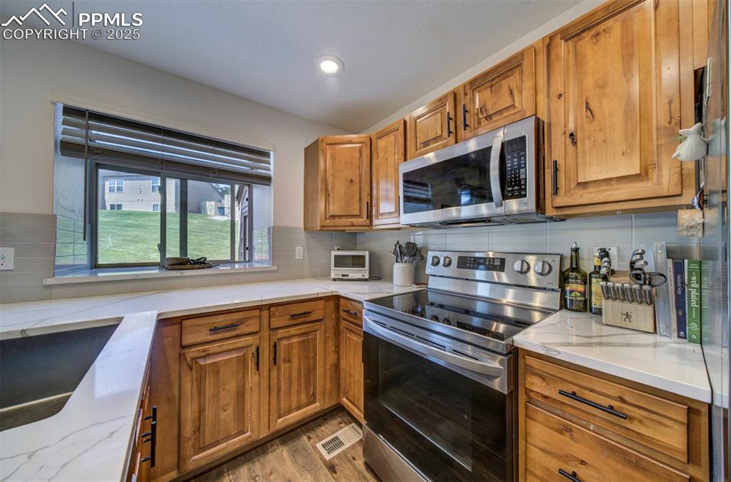 Image 10 of 30: Kitchen with brown cabinetry, stainless steel appliances, light stone count