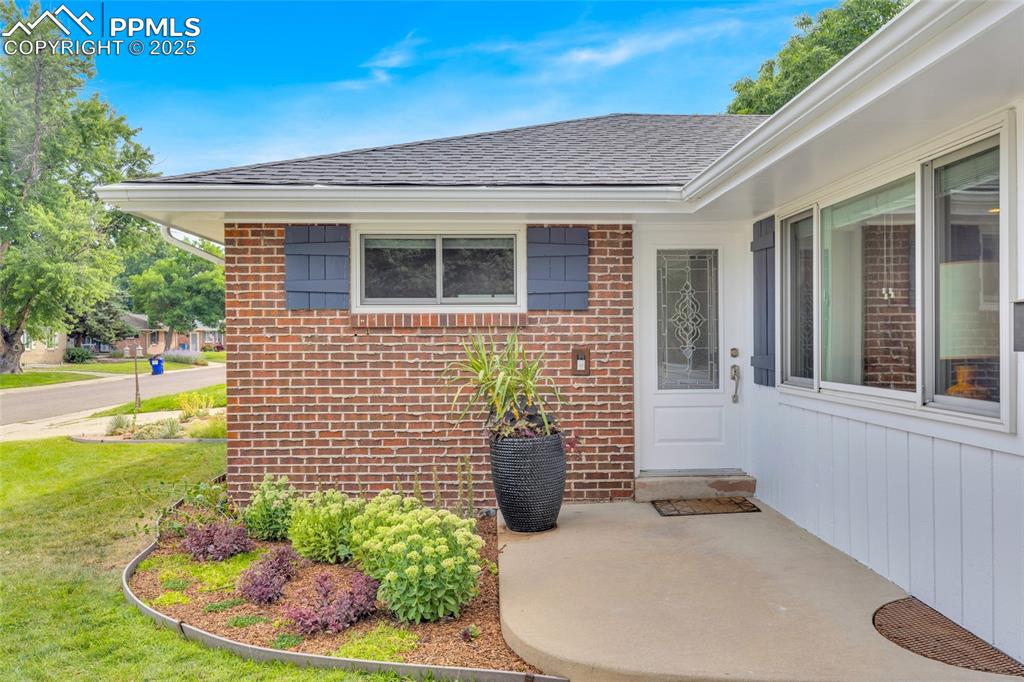 Image 6 of 50: View of exterior entry with brick siding, roof with shingles, and a yard