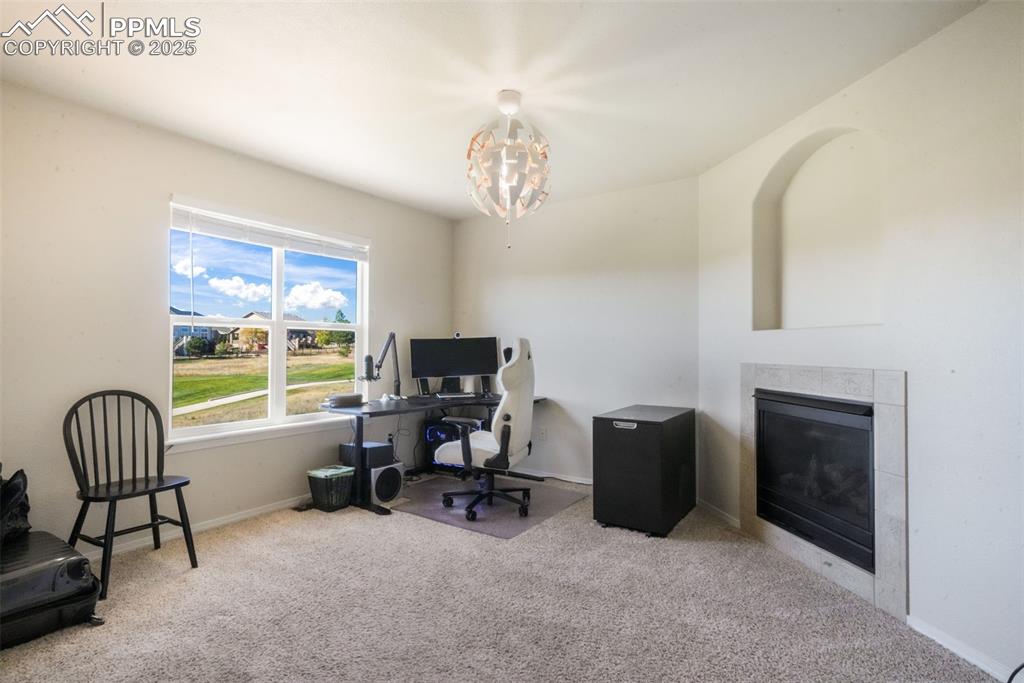 Image 23 of 50: Office space with a tiled fireplace, light colored carpet, and a chandelier