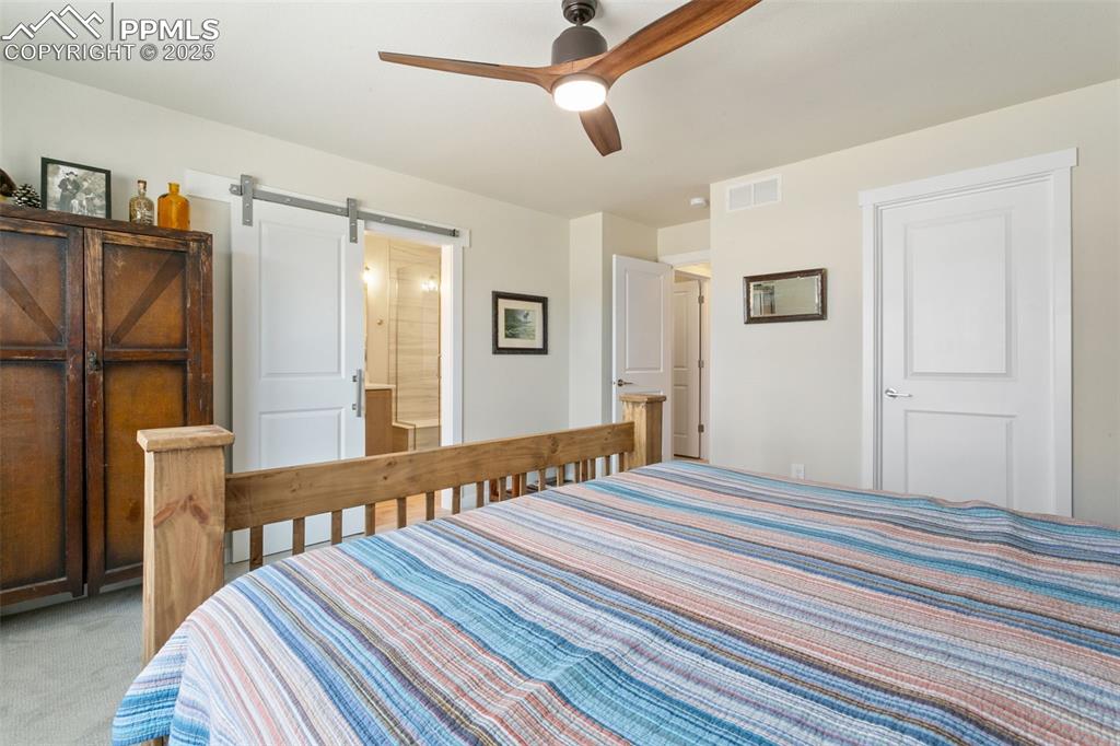 Image 25 of 50: Bedroom with a barn door, light carpet, ensuite bathroom, and a ceiling fan