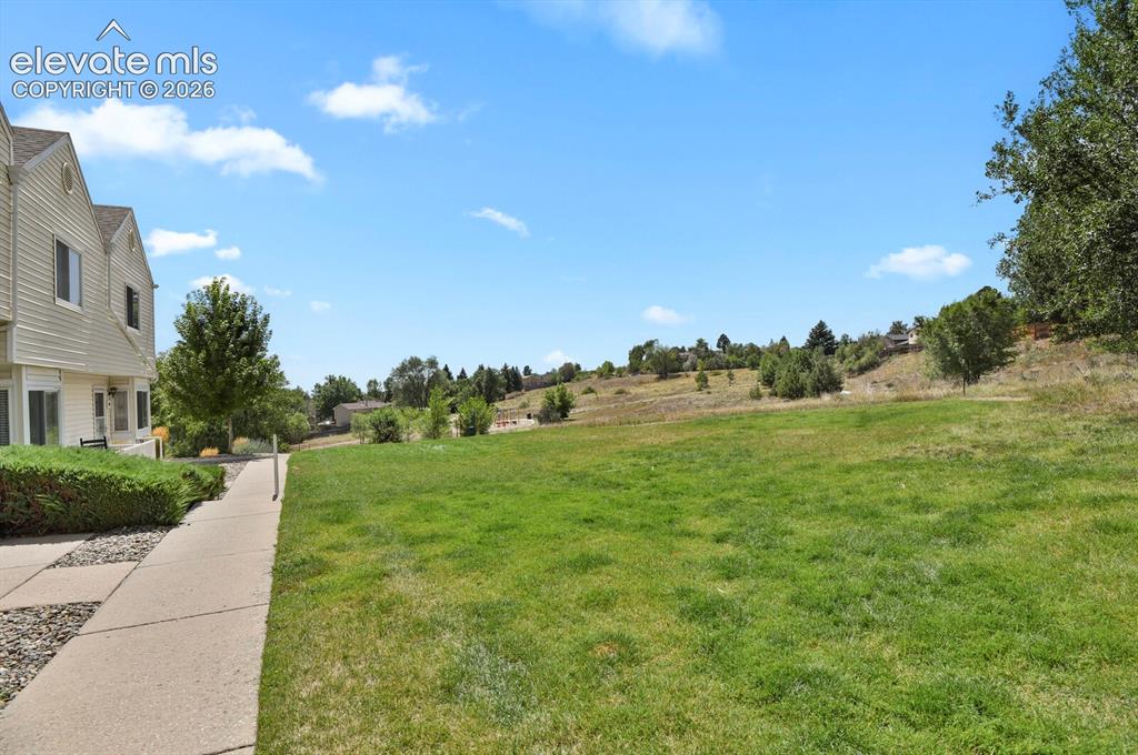 Image 23 of 32: Expansive grassy area with a gentle incline, bordering a paved walkway and 