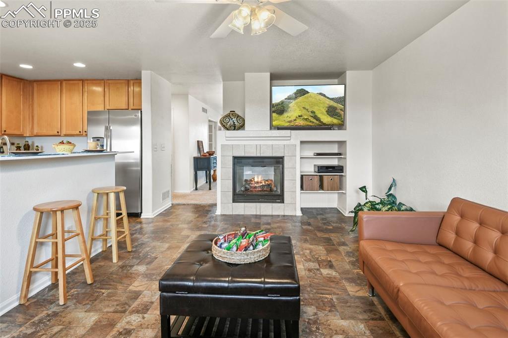 Image 11 of 32: Living room with a tile fireplace, dark stone finish floors, ceiling fan, b
