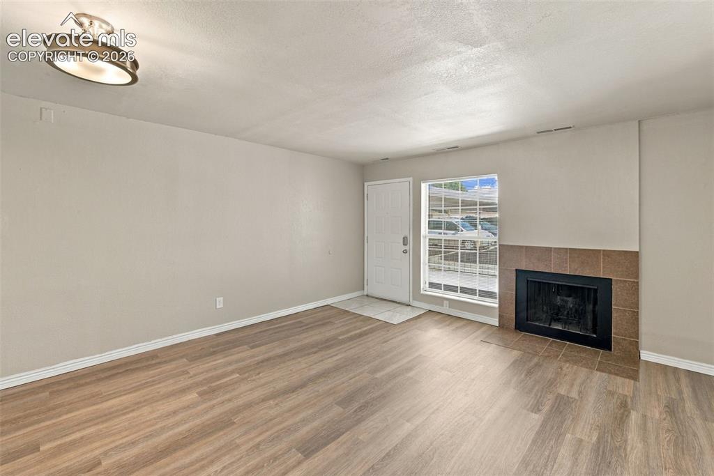 Image 4 of 26: Wood-finish flooring, tiled fireplace with dark surround, white entry door