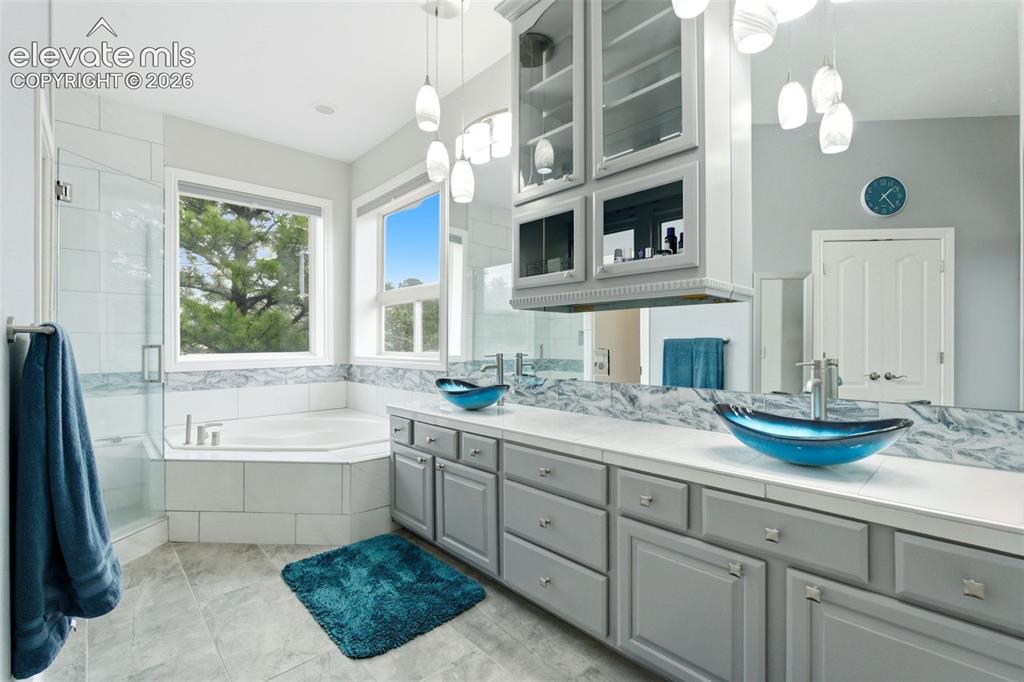 Image 23 of 46: Dual vessel sinks, gray cabinetry, large vanity mirror, soaking tub, and wa