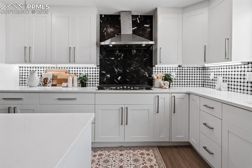 Image 10 of 50: Kitchen with wall chimney range hood, gas cooktop, light countertops, backs