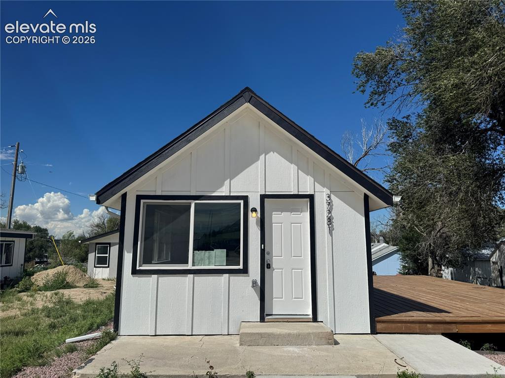 Caption: White vertical panel siding with black trim, black roof, front entry door, and a concrete entry pad