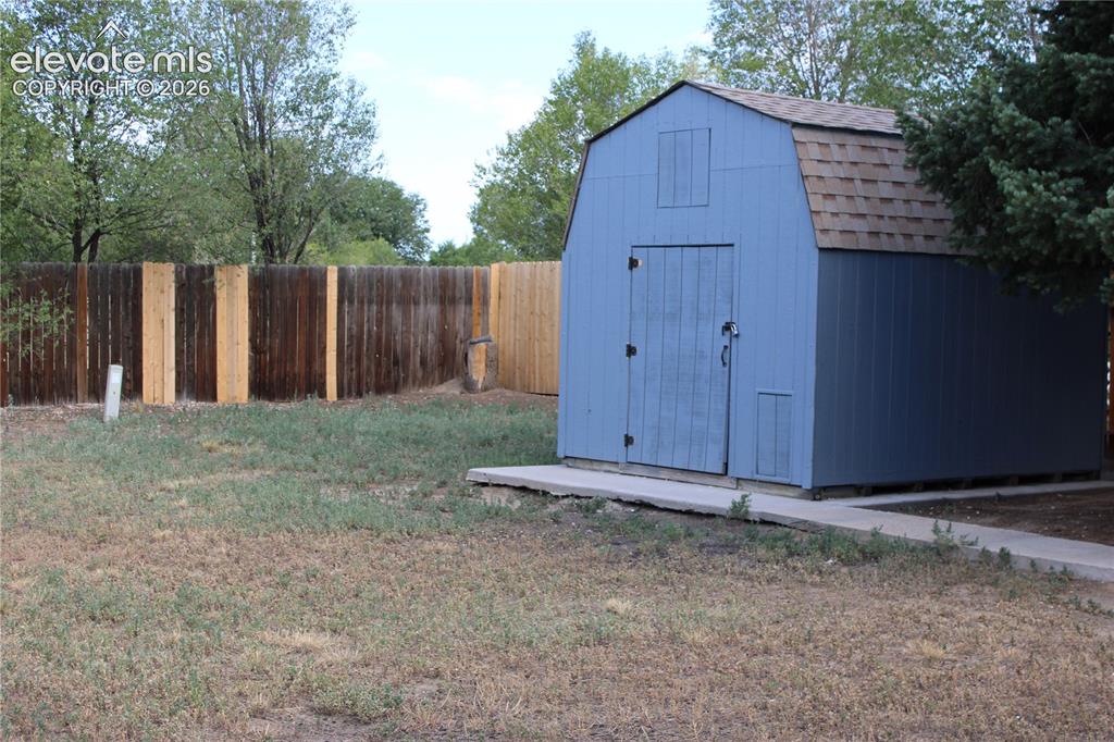 Image 27 of 28: Blue barn-style shed with shingle roofing, wooden privacy fencing, and a co
