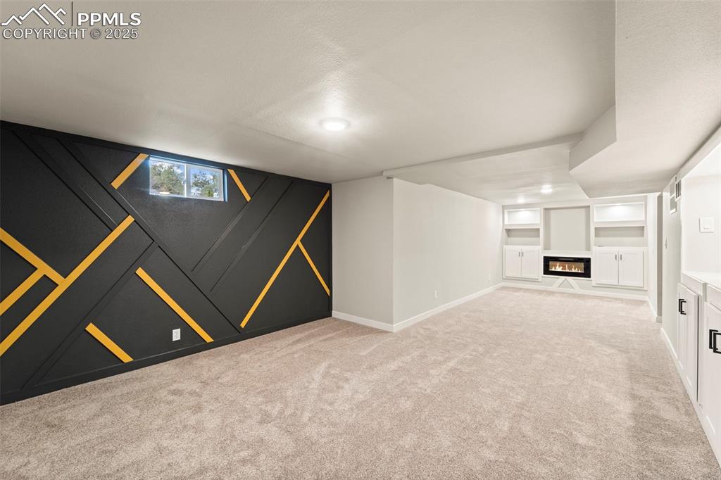 Image 26 of 31: Below grade area with light carpet, built in shelves, a glass covered firep
