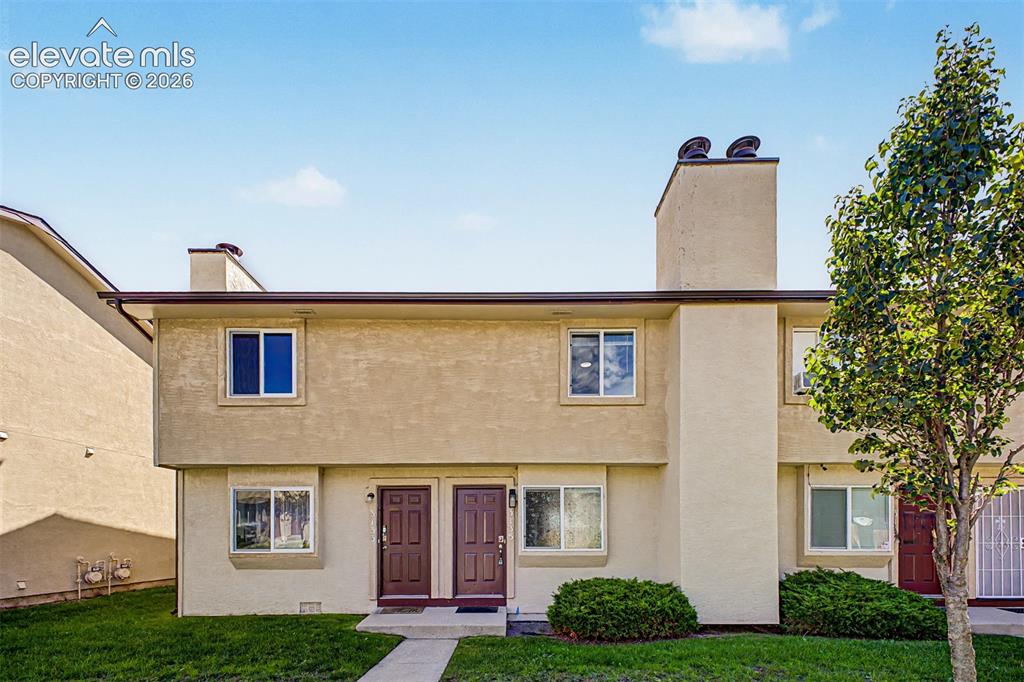 Caption: Stucco exterior with multiple windows, two distinct entry doors, chimney feature, and mature landsca