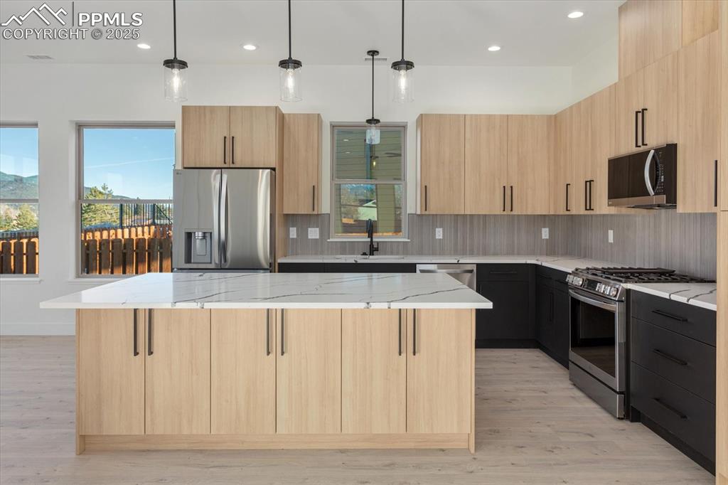 Image 6 of 30: Kitchen featuring light brown cabinets, hanging light fixtures, light stone