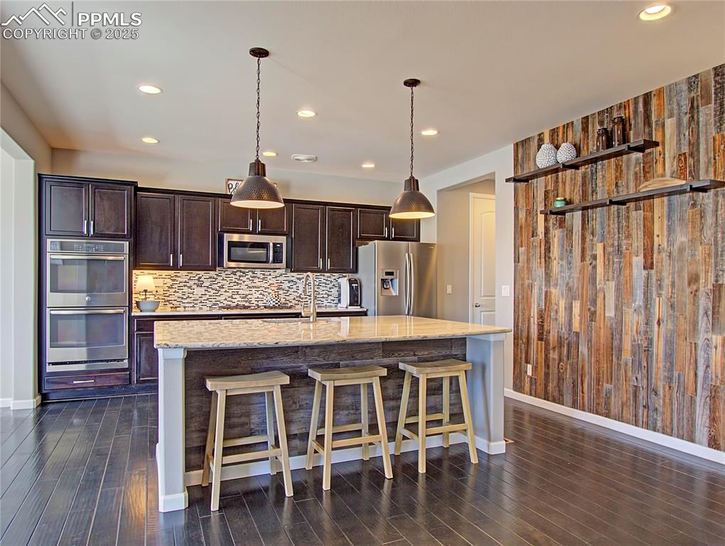 Image 15 of 44: Kitchen featuring dark brown cabinetry, hanging light fixtures, appliances