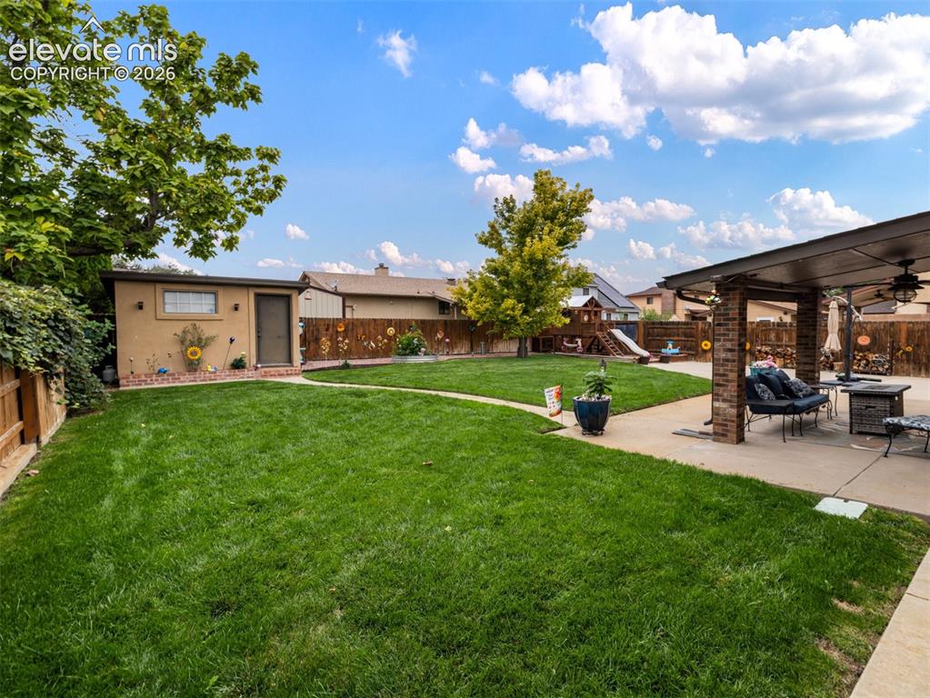 Image 30 of 34: Expansive backyard with a green lawn, covered patio, stucco shed, and wood