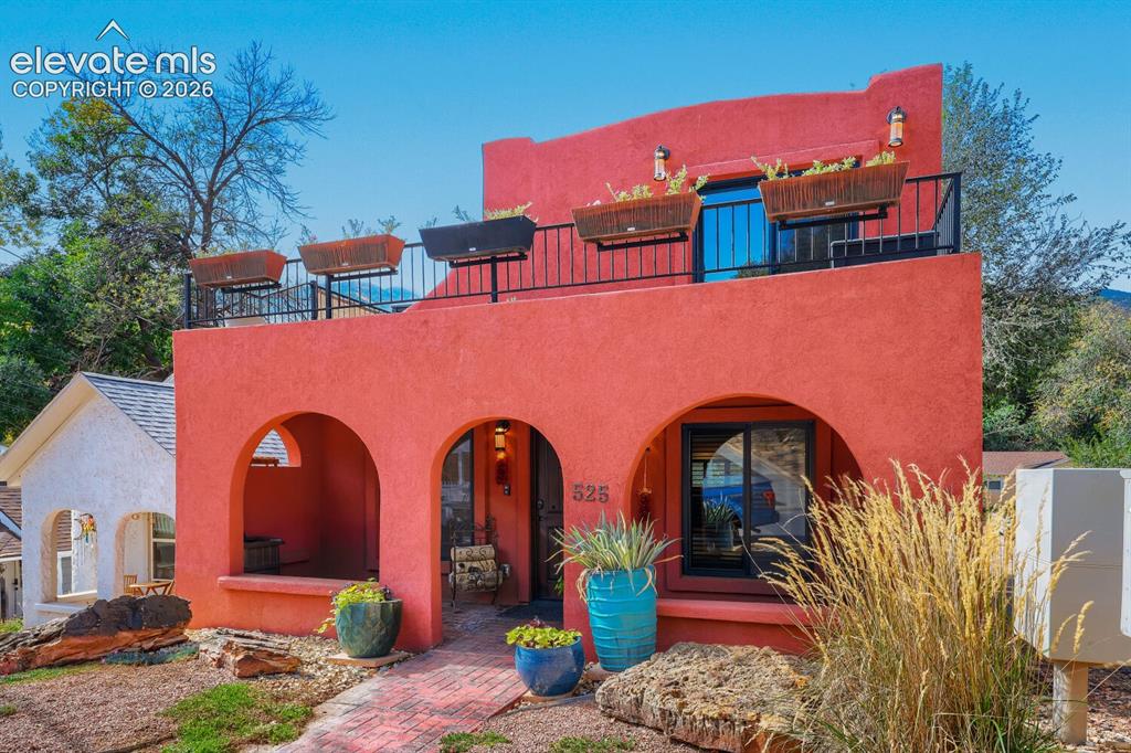 Caption: Terrace with black metal railing, arched entryway, red stucco exterior, black-framed windows, and a 