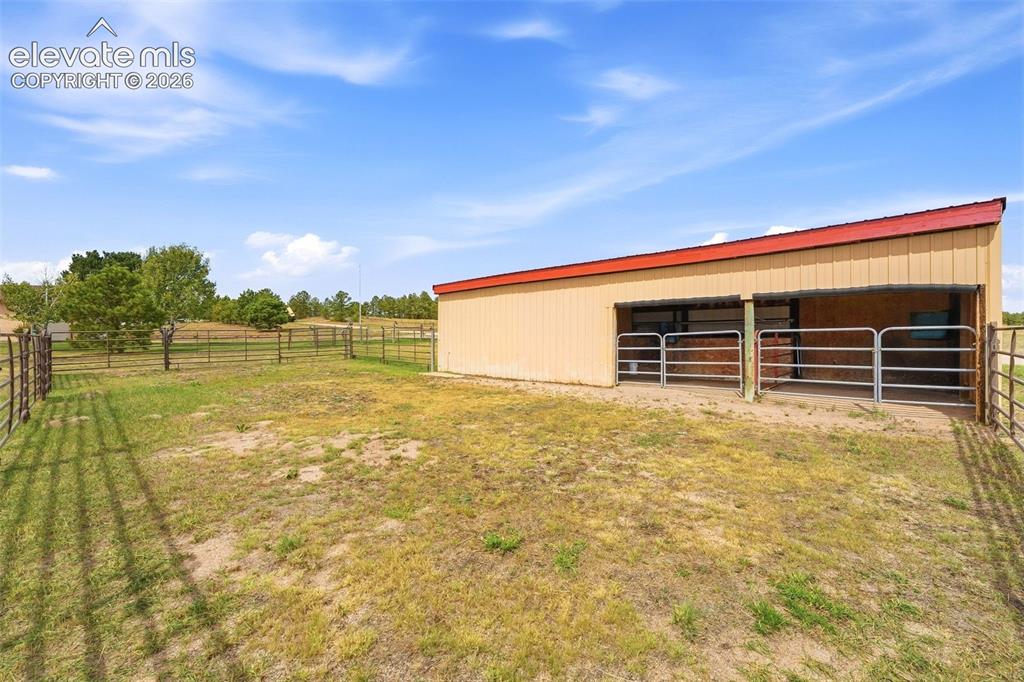 Image 37 of 50: Metal outbuilding with a red sloped roof, two open stalls, and metal fencin