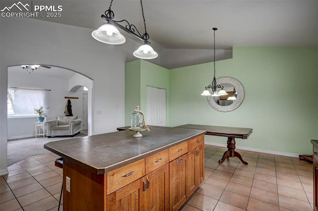Image 15 of 47: Kitchen with brown cabinetry, dark countertops, a center island, pendant li