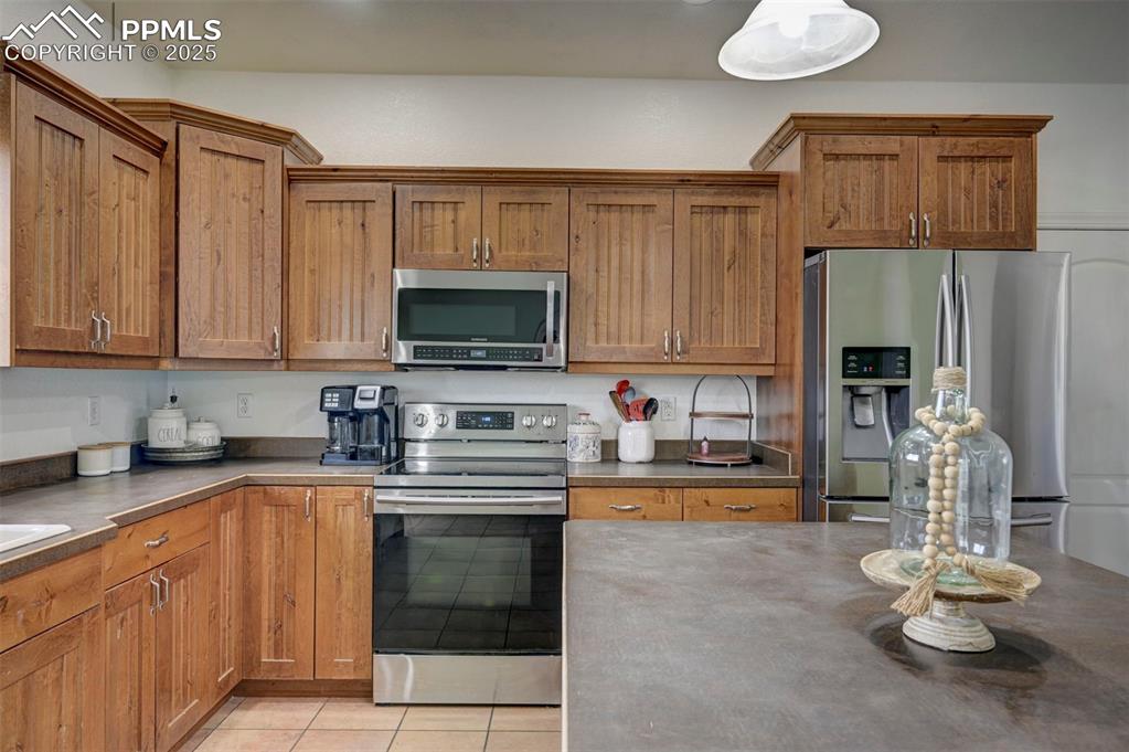 Image 16 of 47: Kitchen with brown cabinets, stainless steel appliances, light tile pattern