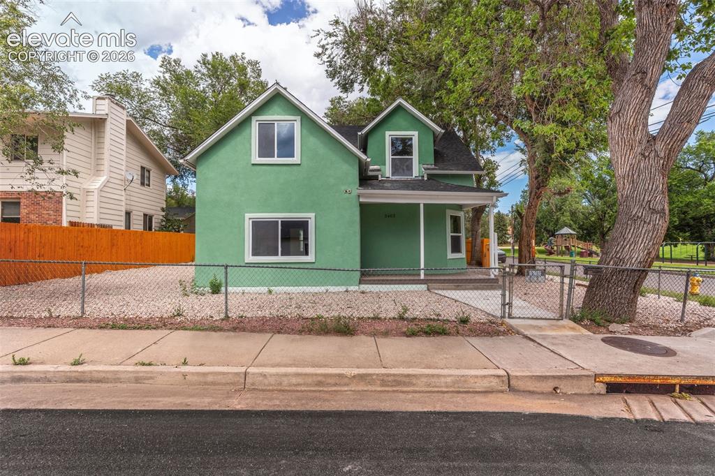 Image 48 of 50: Green stucco exterior with white trim, covered front porch, and chain-link