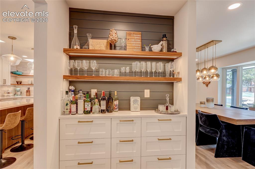 Image 9 of 50: Built-in wet bar with white cabinetry, gold-finish hardware, open wood shel