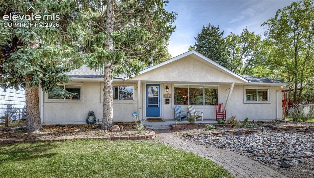 Caption: Stucco exterior, gabled roofline, blue entry door, brick paver walkway, and mature trees